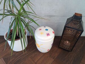 Weisse Urne mit einer Sonne und bunten Blumen bemalt, zwischen einer Pflanze und einer orientalischen Laterne mit brennender Kerze